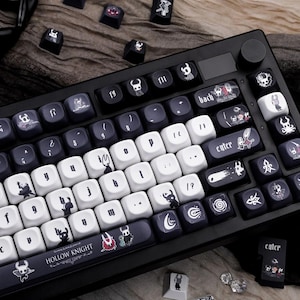 Puede incluir: Teclado mecánico en blanco y negro con teclas personalizadas que presentan arte del juego Hollow Knight. El teclado tiene un marco negro, teclas blancas y gris oscuro, y una perilla de control de volumen. Algunas teclas tienen el texto "enter" y "Hollow Knight".