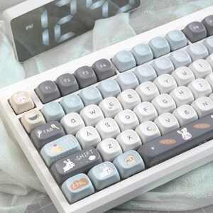 May include: A white mechanical keyboard with light blue, gray, and white keycaps. The keycaps feature various icons and illustrations, including a bunny, a coffee cup, and a cake. The keyboard has a digital clock in the background.