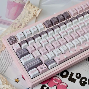 Può includere: Una tastiera meccanica rosa con tasti personalizzati. I tasti sono bianchi, rosa e viola, decorati con cuori, fiocchi e pois. La tastiera ha una cornice rosa chiaro. Il design della tastiera è femminile.