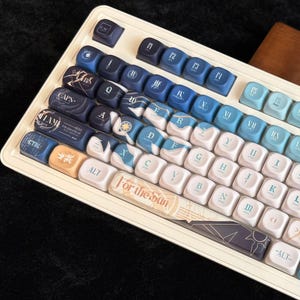 Puede incluir: Un teclado personalizado con base blanca y teclas azules. Las teclas presentan varios diseños, incluyendo temas náuticos, texto y símbolos. El teclado tiene una estética única con una mezcla de colores azul claro y oscuro.