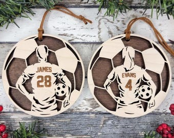 May include: Two wooden soccer ball ornaments with brown leather strings. Each ornament features a soccer player silhouette with a jersey number and name. One reads "JAMES 28" and the other "EVANS 4".