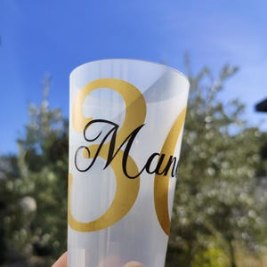 Może przedstawiać: Przezroczysty plastikowy kubek ze złotą liczbą "30" i napisem "Man" czarną czcionką. Kubek jest trzymany na tle błękitnego nieba i zieleni.