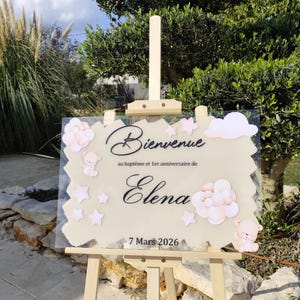 Peut inclure: Panneau de bienvenue transparent sur chevalet en bois. Le panneau présente le mot "Bienvenue" en cursive, ainsi que le nom "Elena" et la date "7 Mars 2026". Des nuages, des étoiles et des ours en peluche roses décorent le panneau.