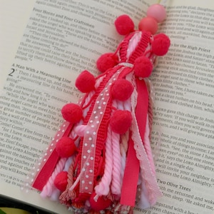 May include: A handmade bookmark with a cluster of red pom-poms, ribbons, and yarn in shades of pink and white. The bookmark is placed on an open book with text visible.