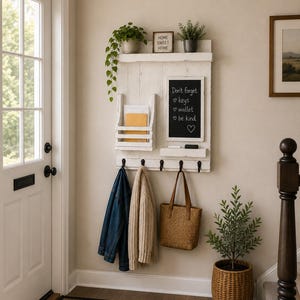May include: A white wooden wall-mounted entryway organizer featuring a mail slot, a chalkboard with the text 'Don't forget: keys, wallet, be kind', a small shelf, and four black metal hooks holding a blue denim jacket, a beige sweater, and a tan tote bag.