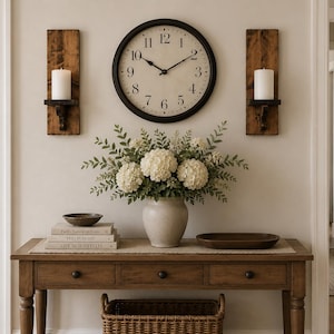 May include: A wooden console table with a wicker basket underneath, topped with a ceramic vase of white hydrangeas, a stack of three books, a small bowl, and a tray. A round black wall clock is flanked by two wooden wall sconces with candles.