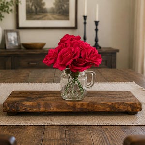 May include: A rectangular dark brown wooden riser with a smooth finish sits on a textured beige placemat. A clear glass mason jar with a handle is placed on the riser, containing a vibrant bouquet of bright red artificial roses with green stems.