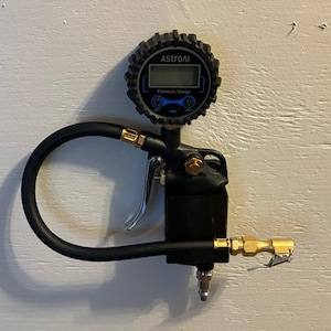 Peut inclure: Manomètre numérique AstroAI noir et argent avec un tuyau noir et une gâchette argentée. La jauge a un affichage numérique et indique "Pressure Gauge". La jauge est montée sur une surface blanche.