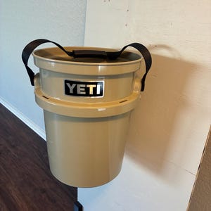 Puede incluir: Cubo YETI de color beige con asas negras. El cubo está colgado en una superficie blanca. El logotipo de YETI se muestra en la parte delantera del cubo.