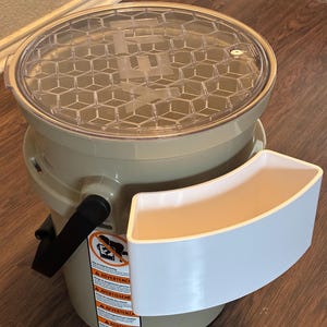 Op de afbeelding: Een beige emmer met een transparante deksel met een honingraatpatroon. Een witte, gebogen bak is aan de zijkant bevestigd. De emmer heeft een zwarte handgreep en een waarschuwingslabel. De deksel heeft de letters "EZ".