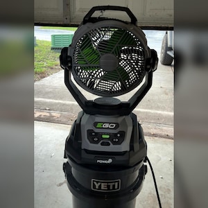 May include: A portable EGO Power+ fan with a gray and black housing and bright green blades. The fan features a digital control panel with plus and minus buttons, a turbo setting, and is mounted on top of a black YETI bucket with a white logo.