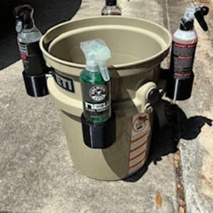 May include: A tan bucket with several spray bottles attached. The bucket has a black handle and the brand name "YETI" is visible. The spray bottles are black and clear, with various colored liquids inside. The image is taken outdoors on a concrete surface.
