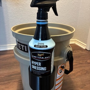 May include: A black spray bottle of Meguiar's Hyper Dressing, held in a black holder attached to a gray Yeti bucket. The bottle has a blue band and the text "D170 READY TO USE". The bucket is on a wooden floor.