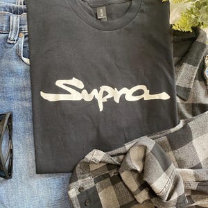 May include: A black t-shirt with the word "Supra" in white script, paired with blue jeans, black sunglasses, a gray plaid shirt, and a silver wristwatch with a blue face. A casual, everyday outfit.