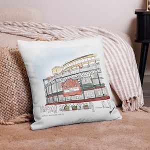 May include: White square throw pillow featuring a watercolor illustration of a baseball stadium. The artwork includes the text "Chicago Cubs" on a red sign, with people and greenery at the base. Signed "Ashley Matselski Art".