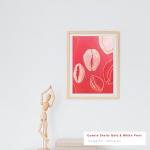 May include: A framed art print featuring a vibrant red background with white and gold cowrie shell illustrations. The artwork is displayed in a light wood frame, adding a natural touch to the design. The print is titled "Cowrie Shells Gold & White Print".