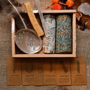 Könnte beinhalten: Eine Holzkiste gefüllt mit spirituellen Gegenständen. Enthält ein weißes Salbei-Bündel, ein Lavendel-Bündel, einen Selenit-Stick, einen Palo Santo-Stick, Truthahnfedern und eine Abalone-Schale. Die Kiste enthält auch kleine Etiketten mit Beschreibungen.