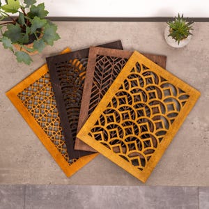 Op de afbeelding: Vier decoratieve houten panelen met ingewikkelde uitgesneden ontwerpen. De panelen hebben geometrische en bladpatronen in bruin- en goudtinten. De panelen zijn gerangschikt op een grijs oppervlak, met kleine potplanten op de achtergrond.