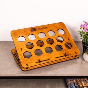 May include: A slanted wooden laptop stand with a honey-brown finish and a series of hexagonal cutouts for ventilation. The top panel features the text ADVANCED CONSULTING LLC printed in black. The stand has a flat base and a front ledge.
