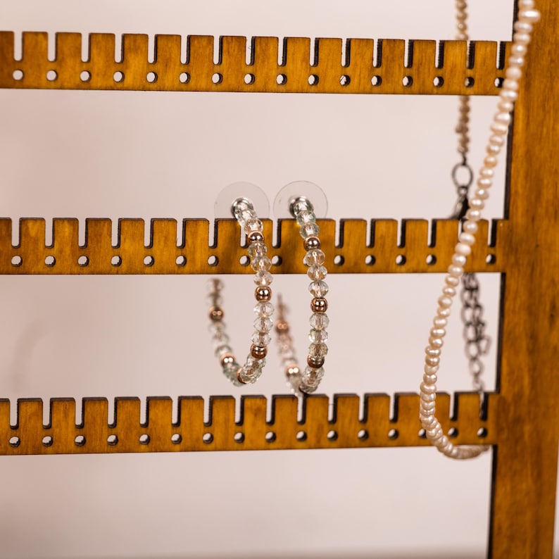 May include: A pair of hoop earrings featuring alternating clear faceted crystal beads and small metallic rose gold-toned spheres. The earrings are displayed on a wooden jewelry stand alongside a strand of small white pearls on a silver chain.