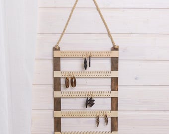 May include: A wall-mounted wooden jewellery organiser featuring five horizontal slats with pre-drilled holes for hanging earrings. The frame is constructed from dark brown wood with lighter natural wood slats and a thick jute rope for hanging.