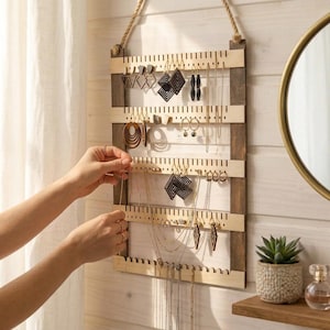 Könnte beinhalten: Ein wandmontierter Schmuck-Organizer aus hellem und dunklem Holz mit einer Seilaufhängung. Der rechteckige Rahmen verfügt über horizontale Leisten mit vertikalen Einkerbungen zum Aufhängen verschiedener Ohrringe, Halsketten und Ketten.