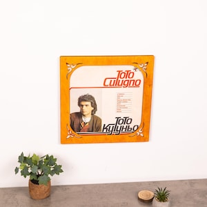 May include: A square wooden frame with decorative corner cutouts containing a vintage vinyl record cover for Toto Cutugno. The cover features a portrait of the artist, the name Toto Cutugno in orange text, and a tracklist in black text.