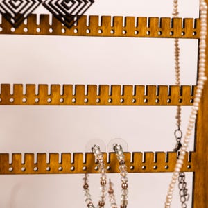 May include: A wooden jewelry display stand with horizontal slats featuring evenly spaced notches for hanging items. The stand holds black geometric earrings, a pair of beaded hoop earrings, and a long strand of small white pearls.
