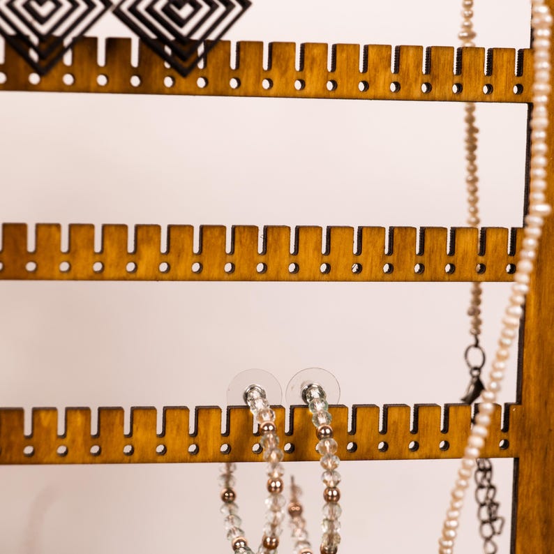 May include: A wooden jewelry display stand with horizontal slats featuring evenly spaced notches for hanging items. The stand holds black geometric earrings, a pair of beaded hoop earrings, and a long strand of small white pearls.