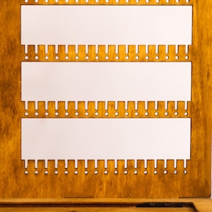 May include: A wooden jewelry display board featuring three horizontal rectangular cutouts. Each cutout is bordered by a series of small, evenly spaced notches and circular holes along the bottom edge, designed for organizing and hanging jewelry items.