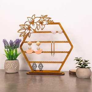 May include: A wooden earring holder in a hexagonal shape, featuring a floral design at the top. The holder is filled with various earrings, including pearl studs, gold hoops, and geometric drop earrings. The holder is displayed on a gray surface.