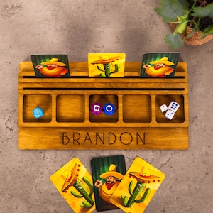 Op de afbeelding: Een houten bordspel-organizer met vijf vierkante vakken en twee horizontale kaartsleuven. Het bord is aan de voorzijde gegraveerd met de naam BRANDON. Het bevat een lichtblauwe dobbelsteen, een paarse dobbelsteen, een blauwe dobbelsteen en drie witte dobbelstenen.
