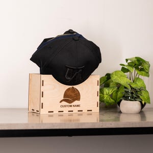 Può includere: Un cappellino da baseball nero con un logo ricamato scuro appoggiato su una scatola espositiva in legno chiaro. La scatola presenta un'incisione laser di un cappellino sopra il testo CUSTOM NAME, con bordi dentellati e forma rettangolare.