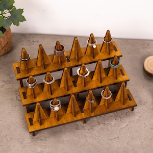 May include: A three-tiered wooden ring display stand with multiple rings on display. The stand is made of light brown wood and features triangular supports. Various rings with different designs are showcased.