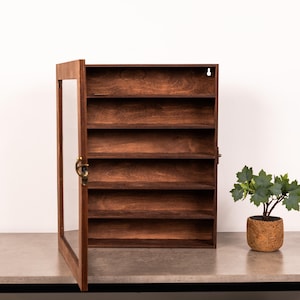 Könnte beinhalten: Ein dunkel gebeizter Holz-Vitrinenschrank mit Glastür und mehreren Regalen. Die Tür ist geöffnet und gibt den Blick ins Innere frei. Rechts steht eine kleine Topfpflanze. Der Schrank steht auf einer grauen Oberfläche.