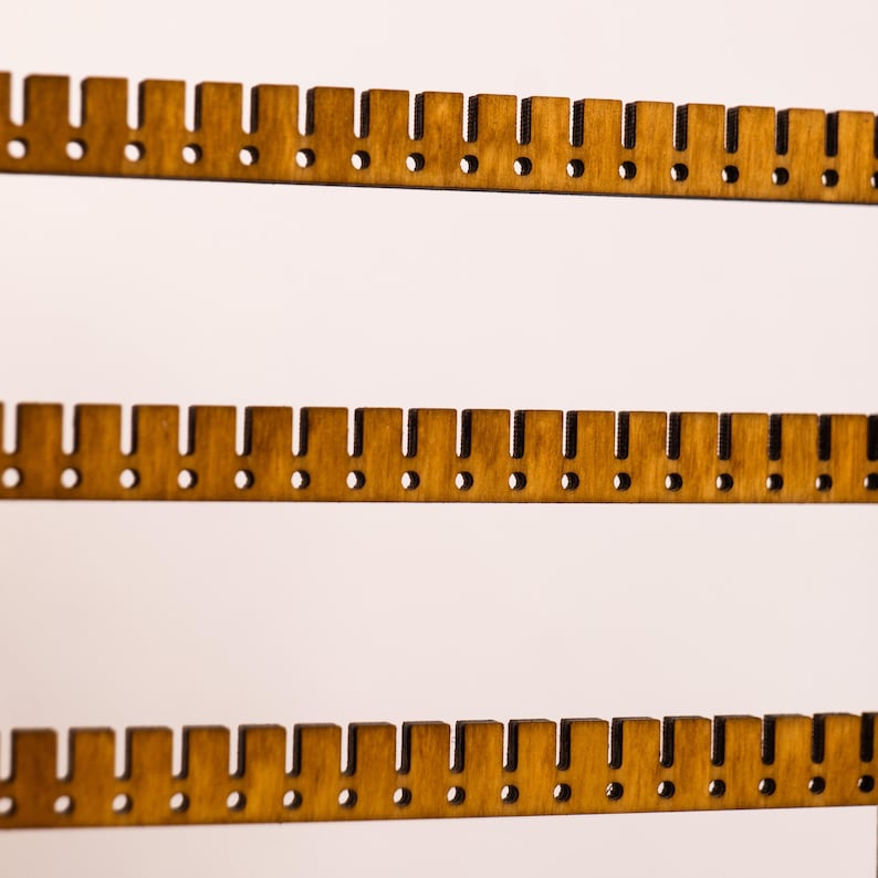 May include: A light brown wooden jewelry display rack featuring three horizontal bars. Each bar has a series of evenly spaced vertical notches along the top edge and a corresponding row of small circular holes positioned below each notch.