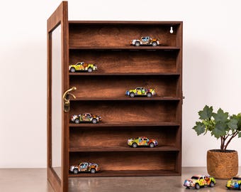 Puede incluir: Una vitrina de madera teñida oscura con puerta de cristal, que muestra varios camiones de juguete coloridos en sus estantes. La vitrina tiene un tirador color latón y está sobre una superficie gris, con una planta en maceta a la derecha.