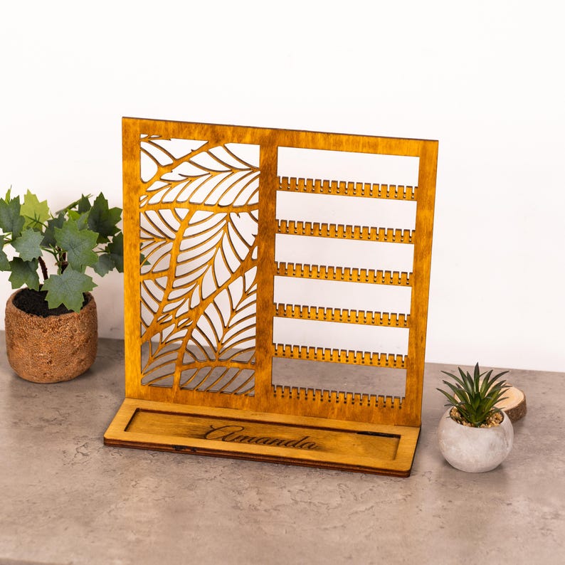 May include: A rectangular wooden jewelry display stand with a warm brown finish. The left side features a laser-cut leaf pattern, while the right side has six horizontal rows with small notches for hanging earrings. The base is engraved with the name Amanda.
