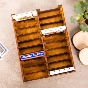 Puede incluir: Un soporte de madera para cartas, de color marrón cálido, con múltiples ranuras que contienen cartas. Se ve una caja de cartas "PLASTIC COATED". El soporte está diseñado para organizar y exhibir naipes.