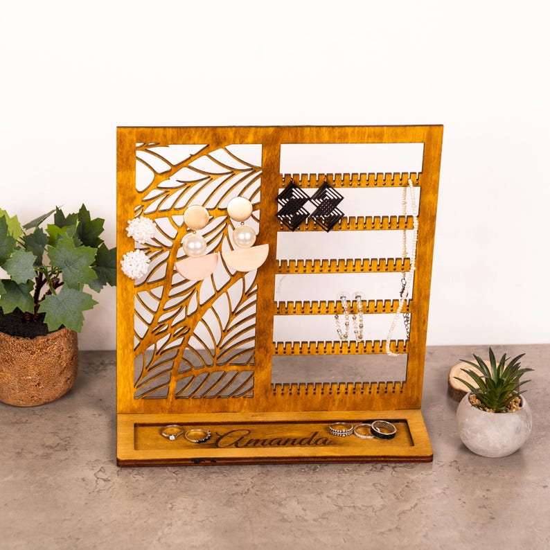 May include: A wooden jewelry organizer with a natural finish, featuring a left panel with a laser-cut leaf pattern and a right panel with horizontal notched bars for earrings. The base includes a tray with the engraved name Amanda and several rings.