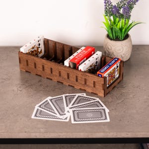 May include: A dark brown wooden playing card holder containing several decks of cards. The holder has slots for each deck. Some decks are red and white, and one is labelled "PLASTIC COATED". A fan of playing cards is in front of the holder.