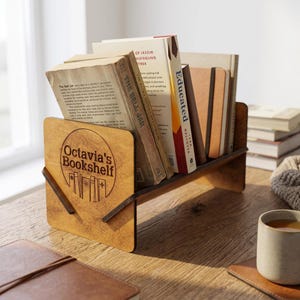 以下が含まれることがあります： Octavia's Bookshelfという文字と本のグラフィックがレーザー刻印された円形ロゴ付きの木製デスクトップブックラック。ラックには『The Bell Jar』や『Educated』などのハードカバー本が数冊、水平に並べて収納されています。