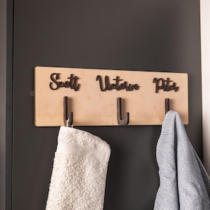 May include: A wooden wall-mounted coat rack with three dark metal hooks. The names "Scott", "Victoria", and "Peter" are cut out above each hook. A white towel and a blue patterned towel hang from the hooks.