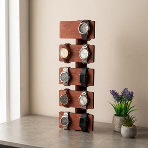 Puede incluir: Un expositor de relojes de madera con seis relojes. El soporte es de madera marrón oscuro y tiene plataformas horizontales para sujetar los relojes. Los relojes tienen correas de varios colores, incluyendo marrón, plateado y negro.