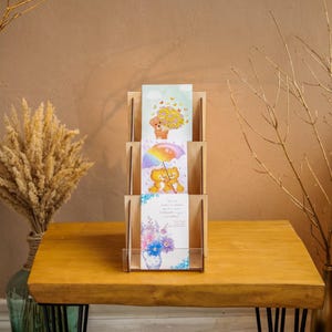 Op de afbeelding: Een houten displaystandaard voor wenskaarten met drie sleuven, elk met een andere geïllustreerde kaart. De kaarten bevatten afbeeldingen van teddyberen, bloemen en een regenboogparaplu. De standaard staat op een houten tafel.