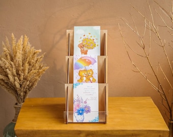 Pode incluir: Um expositor de madeira de três níveis com três cartões de felicitações retangulares. O cartão superior mostra um urso com flores amarelas, o do meio dois ursos sob um guarda-chuva arco-íris e o inferior um vaso azul com flores.