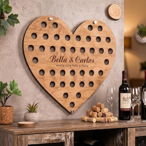 May include: A heart-shaped wooden wine cork holder featuring numerous circular cutouts for cork storage. The center is engraved with the names Bella & Carlos and the text every cork tells a story. It is mounted with silver hardware.