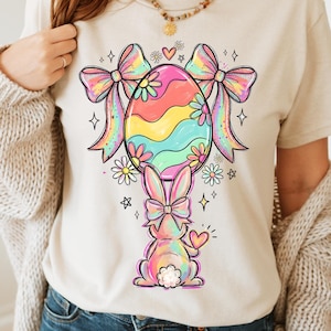 Peut inclure: T-shirt beige avec un motif de Pâques coloré. Le design comprend un œuf rayé arc-en-ciel, des nœuds et un lapin, ainsi que des fleurs, des étoiles et un cœur. Un vêtement décontracté pour tous les jours.
