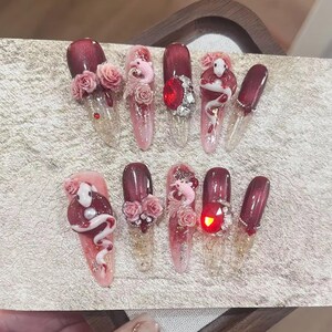 May include: A collection of ten long, artificial nails with diverse designs. The nails display a pink to burgundy gradient, adorned with 3D roses, rhinestones, and sculpted snake figures. Some nails have a clear base, showcasing intricate nail art.