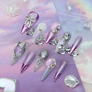 May include: An assortment of artificial nails in various designs and colors. The nails feature a range of embellishments, including bows, pearls, and metallic accents. The color palette includes shades of purple, silver, and white.